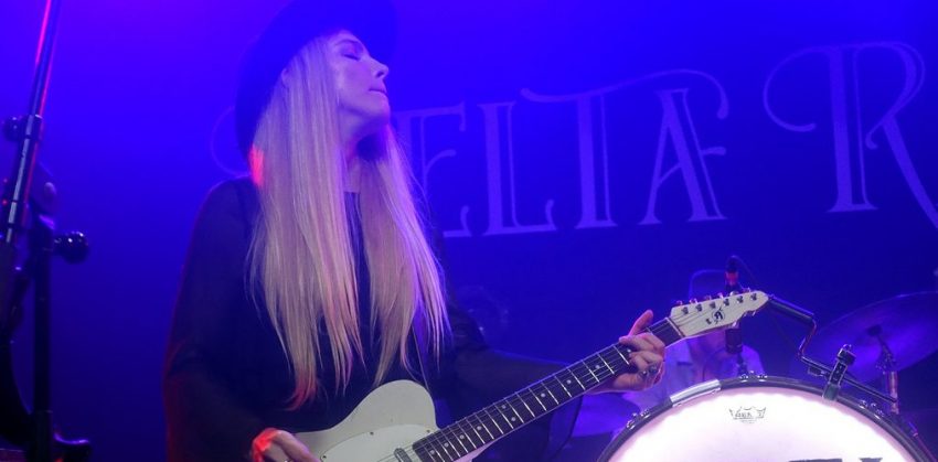 Delta Rae band member Brittany Holljes standing with a guitar next to the band logo.